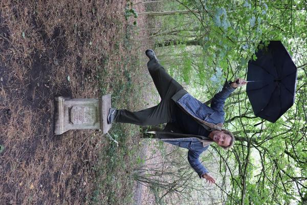 A man in a blue jacket playfully balances on a weathered stone pedestal in a wooded area, holding a black umbrella